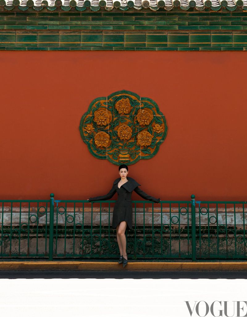 Adult female fashion model in a black dress poses against an orange wall with green architectural features in an outdoor setting.