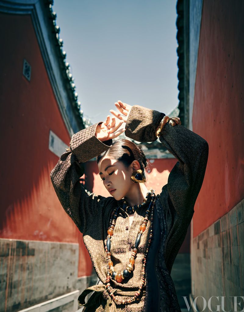 An Asian woman poses in a courtyard wearing a brown textured dress and a large beaded necklace with her hands above her head.