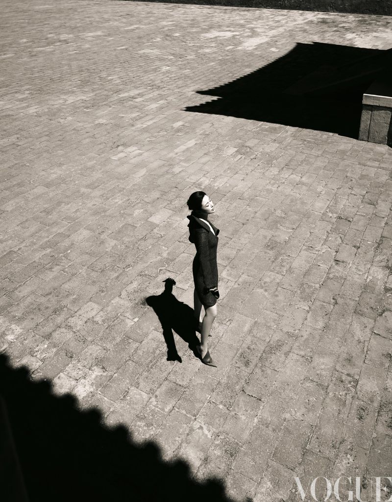 Fashion editorial shot from above featuring an Asian woman in a dark coat standing in a courtyard with stone tiles and shadows.