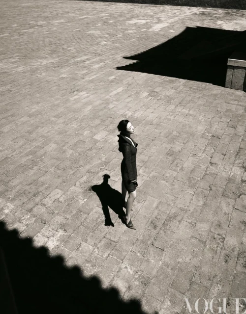 Fashion editorial shot from above featuring an Asian woman in a dark coat standing in a courtyard with stone tiles and shadows.