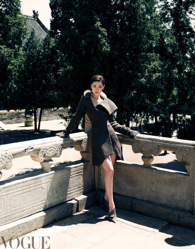 Woman posing in a stylish coat on a stone balcony with trees and a building visible in the background, Vogue logo in the bottom left.