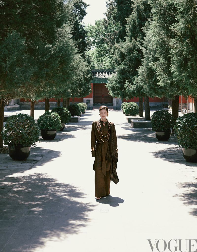 Fashion editorial photo features a woman in a brown dress, walking through a courtyard lined with trees in an outdoor setting on a sunny day.
