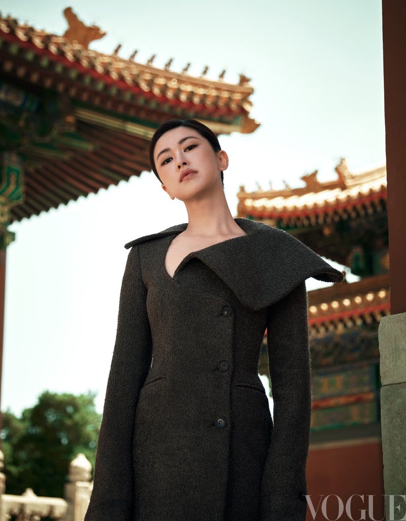 Adult female model wears a dark grey coat with a wide collar while posing in front of a red traditional Chinese building and trees.