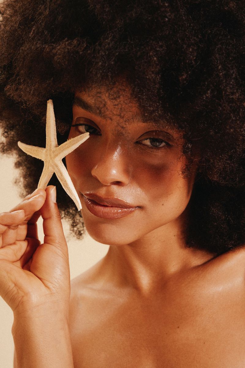 An adult female poses in a studio with a starfish covering her eye, creating a summery, tropical mood with minimal makeup and lighting.