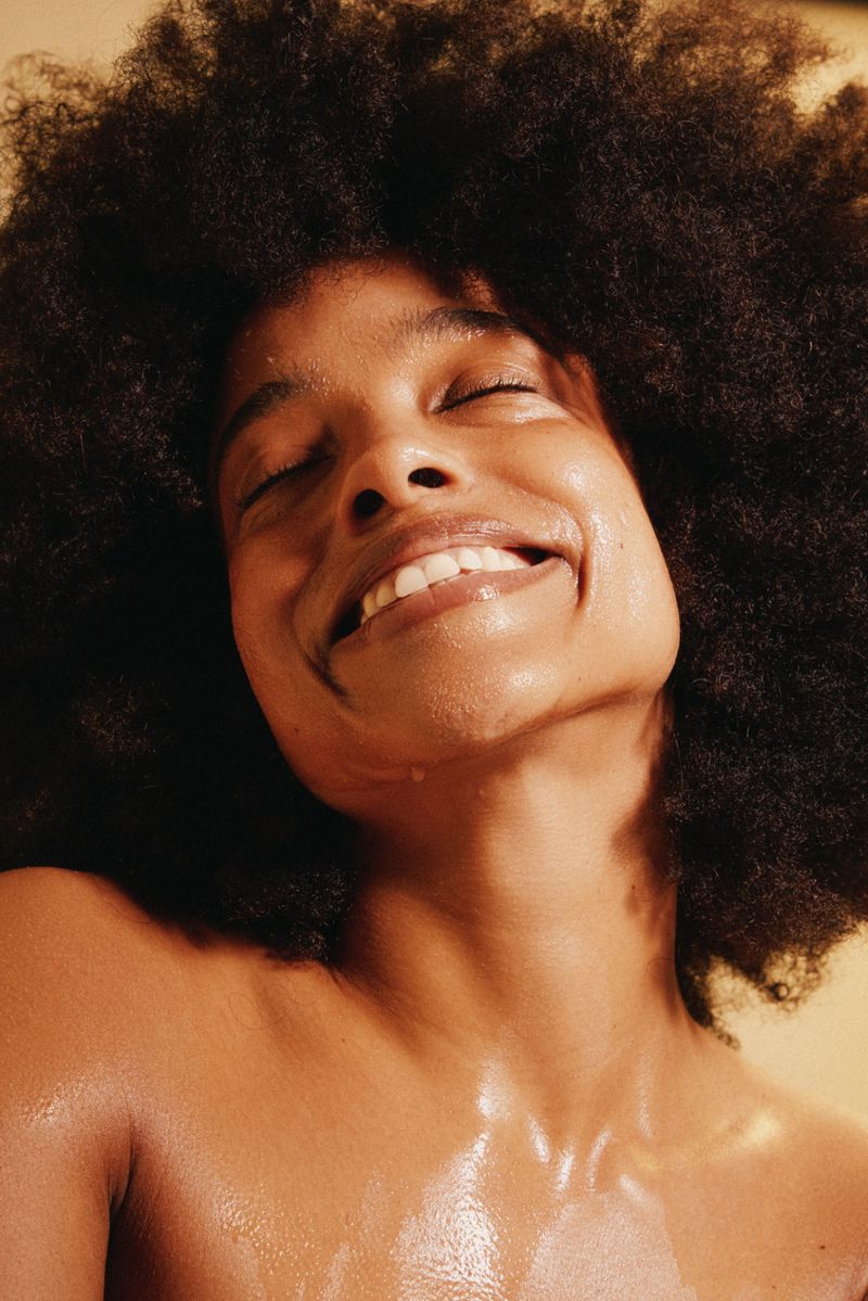 Close-up portrait of a smiling woman with dark curly hair and moist skin against a peach backdrop in a studio setting. Beauty shot.