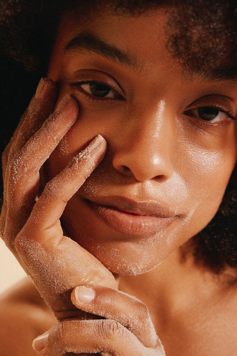 Close-up beauty portrait of a Black woman with dewy, glowing skin resting sand-dusted hands along her cheek and jawline.