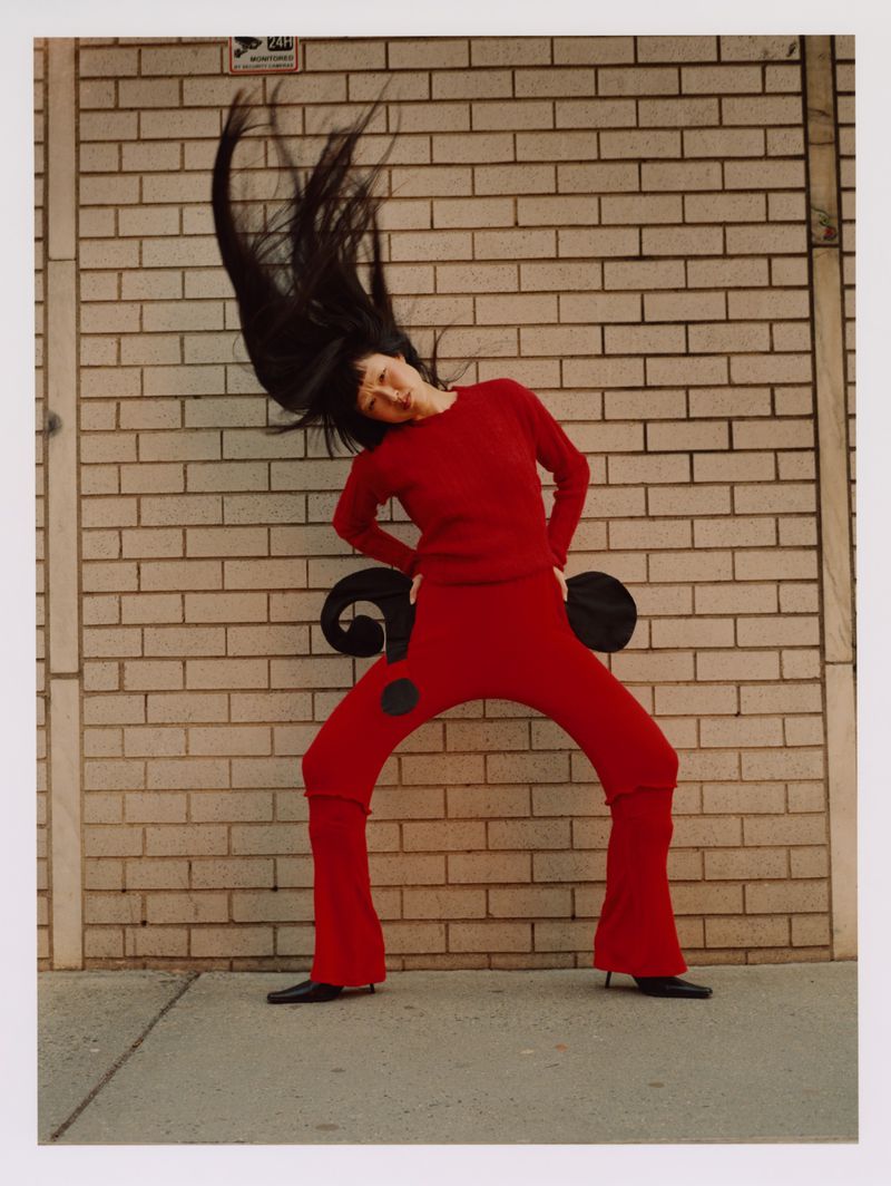 A woman in red attire strikes a dynamic pose outdoors against a brick wall, her dark hair flying, capturing a fashion-forward moment.