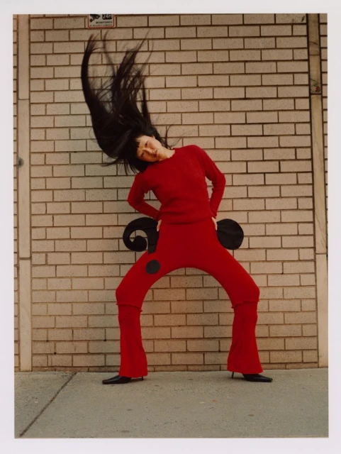 A woman in red attire strikes a dynamic pose outdoors against a brick wall, her dark hair flying, capturing a fashion-forward moment.