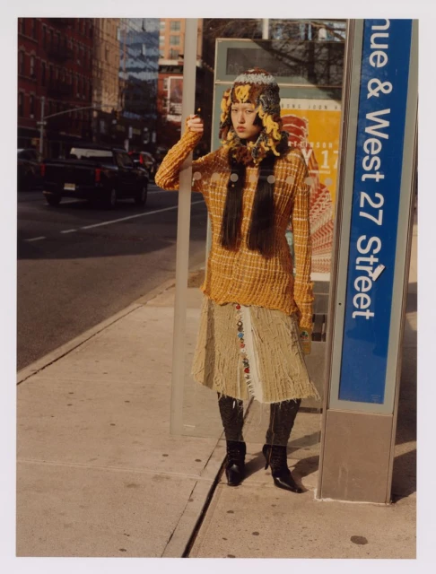 A fashionable woman in a patterned sweater and hat stands on a city sidewalk next to a bus stop sign on a sunny day in an urban setting.