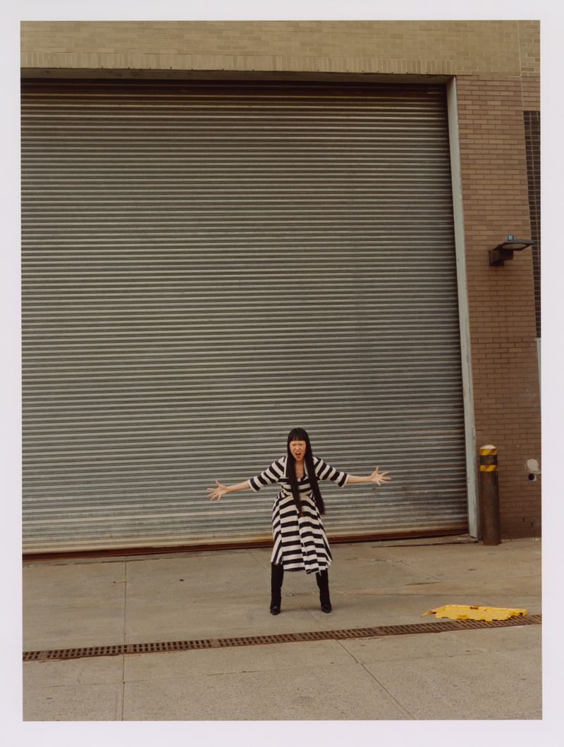 Full-length shot of a person with black hair and bangs standing in a striped dress with arms outstretched in front of a grey metal door.