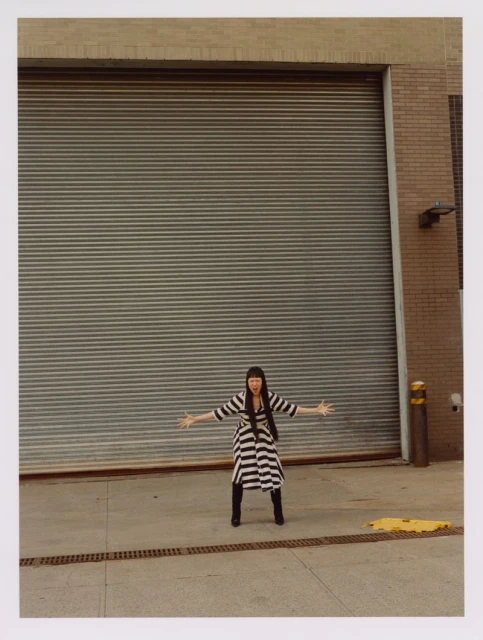 Full-length shot of a person with black hair and bangs standing in a striped dress with arms outstretched in front of a grey metal door.