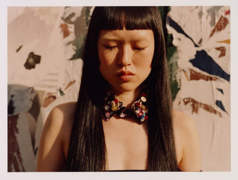 Asian woman with long black hair and a colorful beaded collar posing in front of a weathered wall with torn posters in soft light.