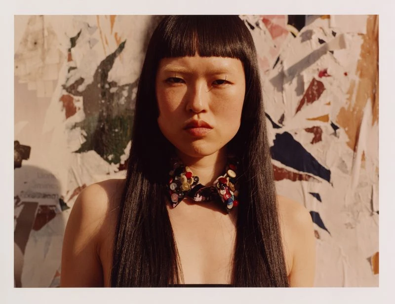 Close-up of an Asian woman with blunt bangs and straight dark hair wearing an embellished bow necklace against a distressed wall.