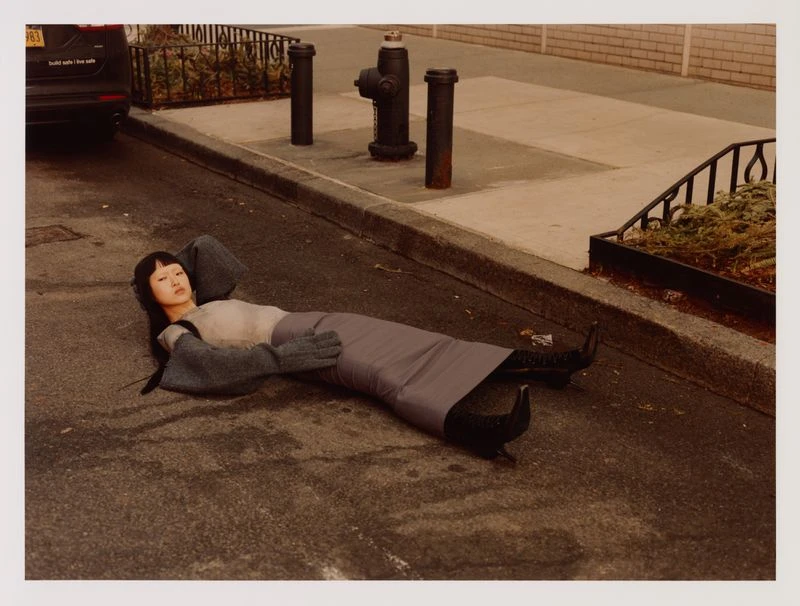 Woman in grey dress and sleeves lies on a city street next to fire hydrant. A black SUV with white text drives by in background.