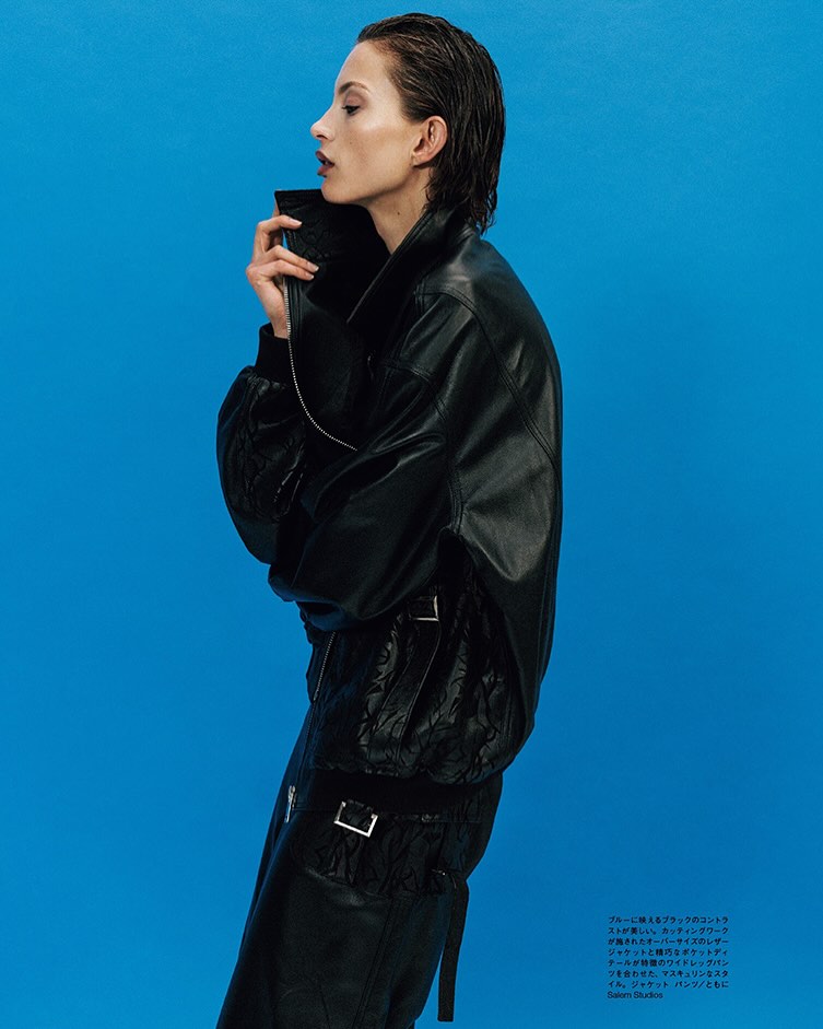 Fashion shot: Woman in profile against a blue backdrop, wearing a black leather jacket and skirt, with dark, slicked-back hair.
