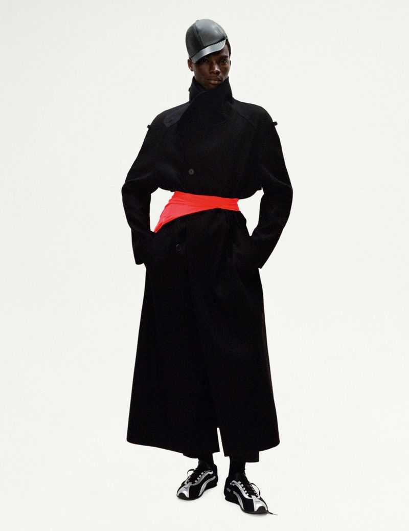 An adult Black male model in a black coat with a red sash, black cap, and sneakers, posing in a photo studio against a white backdrop.