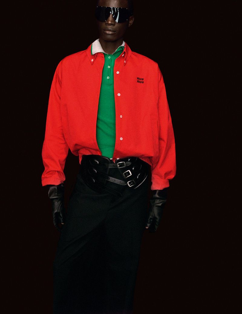 An androgynous dark-skinned person wears a red button-down shirt, green polo shirt, black skirt, black gloves, and sunglasses against black backdrop.