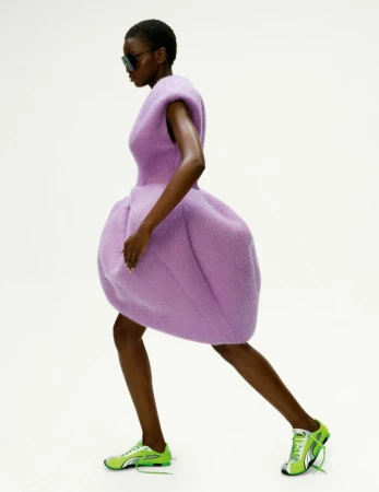 Fashion editorial shot of a slender, dark-skinned woman in a lilac sculptural dress and lime green sneakers posing against a stark white backdrop.