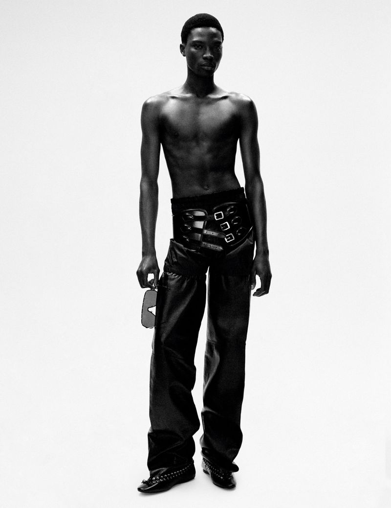 Black and white fashion portrait of a shirtless Black male model wearing studded leather pants and holding sunglasses against a white background.