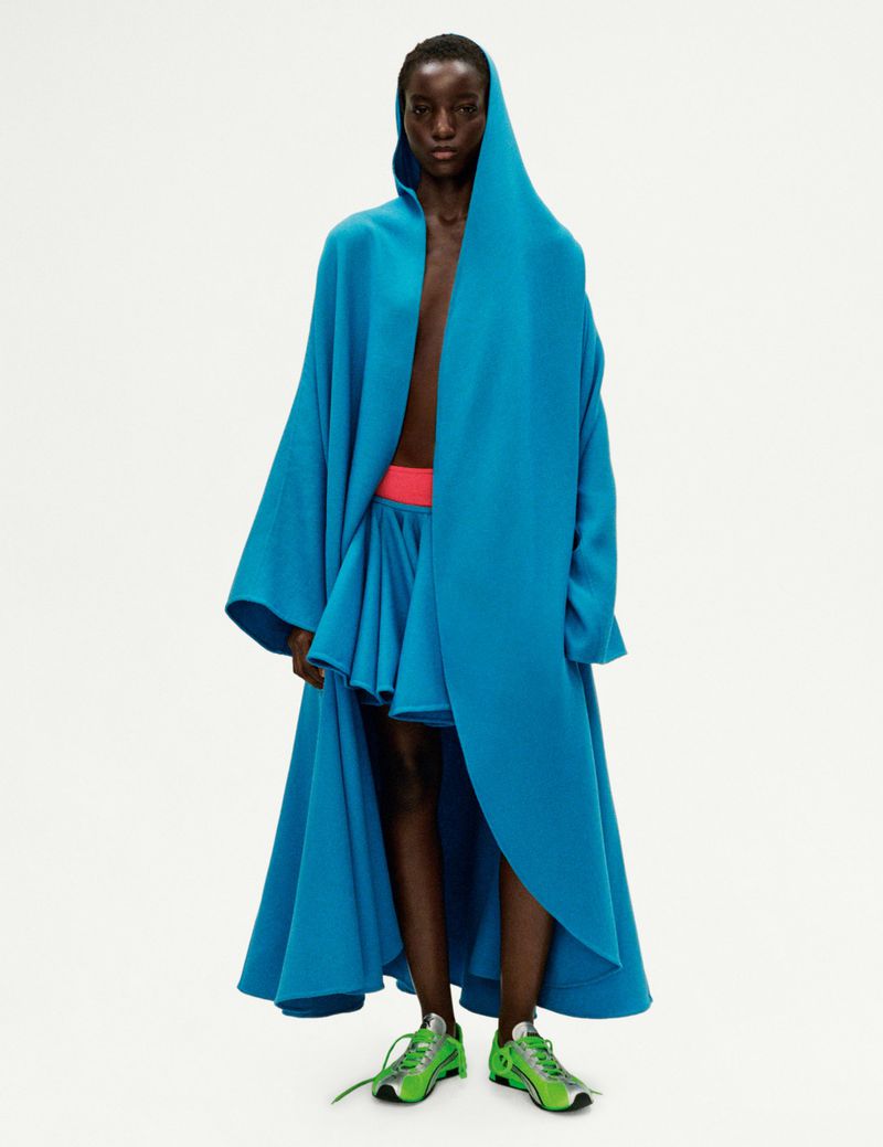 An adult Black female model wearing a hooded blue cape and matching skirt poses in a studio setting with bright green sneakers.