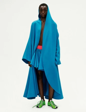 An adult Black female model wearing a hooded blue cape and matching skirt poses in a studio setting with bright green sneakers.