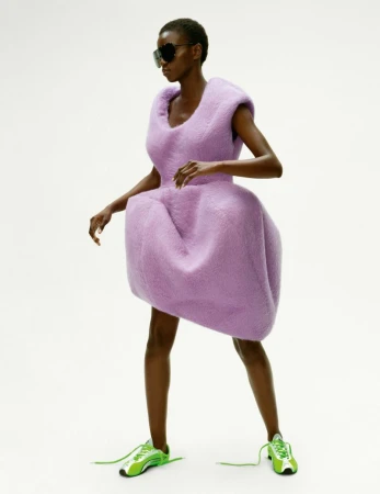 An African American woman models a lavender textured dress with green sneakers against a white background, wearing dark sunglasses.