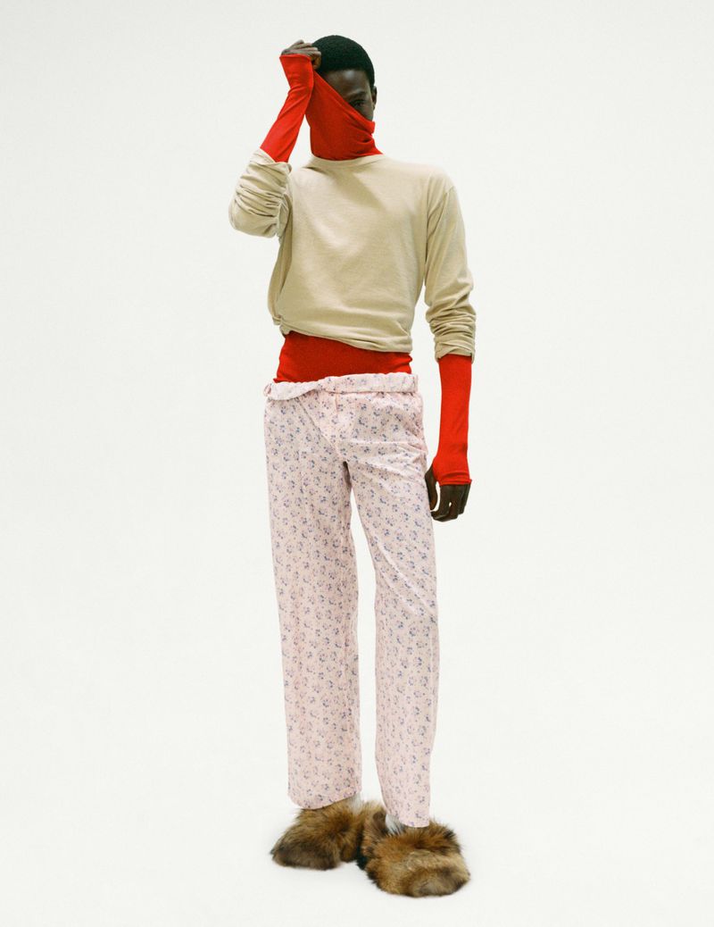 Dark-skinned male model in red and beige separates with patterned pink pants and fur slippers posing in front of a white backdrop.