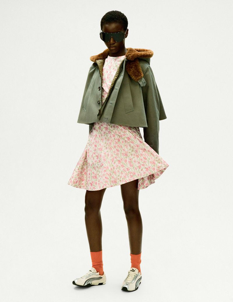 Woman in a floral dress, jacket with a fur collar, and sneakers stands in a brightly lit studio setting, creating a fashion-forward, stylish look.