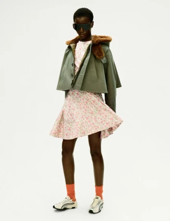 Woman in a floral dress, jacket with a fur collar, and sneakers stands in a brightly lit studio setting, creating a fashion-forward, stylish look.