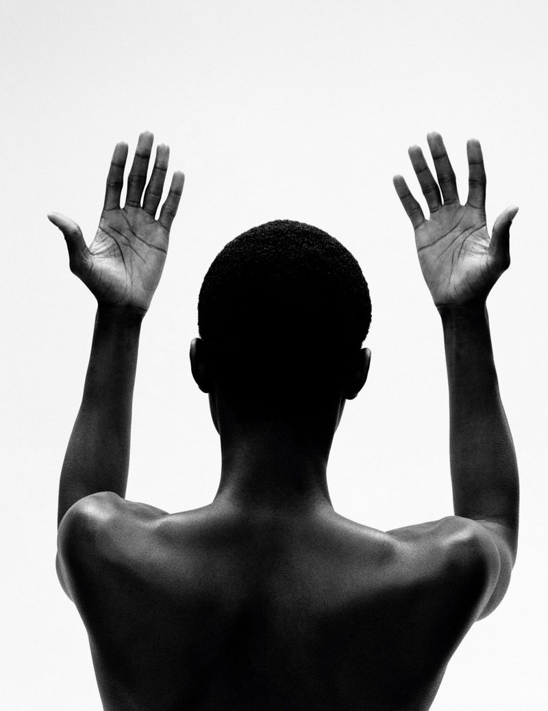 Back view of an African American adult with hands raised against a white background in a high-contrast, black and white photograph.