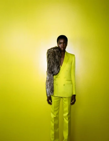 Fashionable black model in a bright yellow suit with a fur stole poses against a solid yellow background in a studio shot.