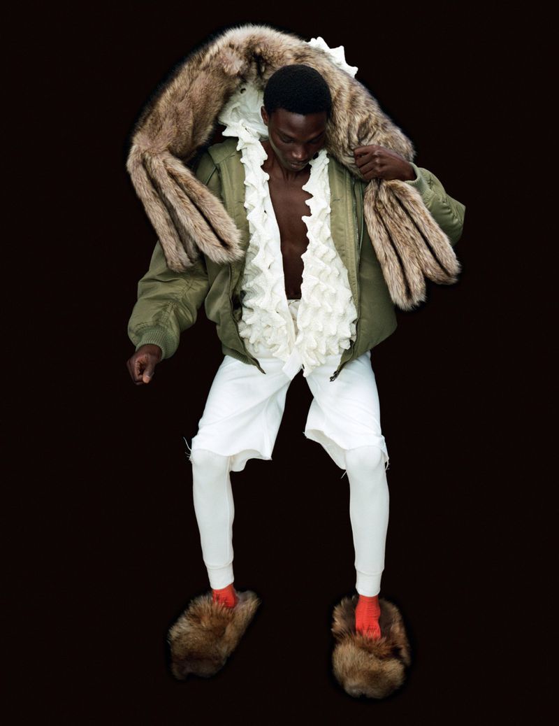Fashion shot of a Black male wearing a green bomber jacket, white shorts and leggings, large furry slides, and a fur stole against a black backdrop.