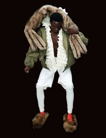 Fashion shot of a Black male wearing a green bomber jacket, white shorts and leggings, large furry slides, and a fur stole against a black backdrop.