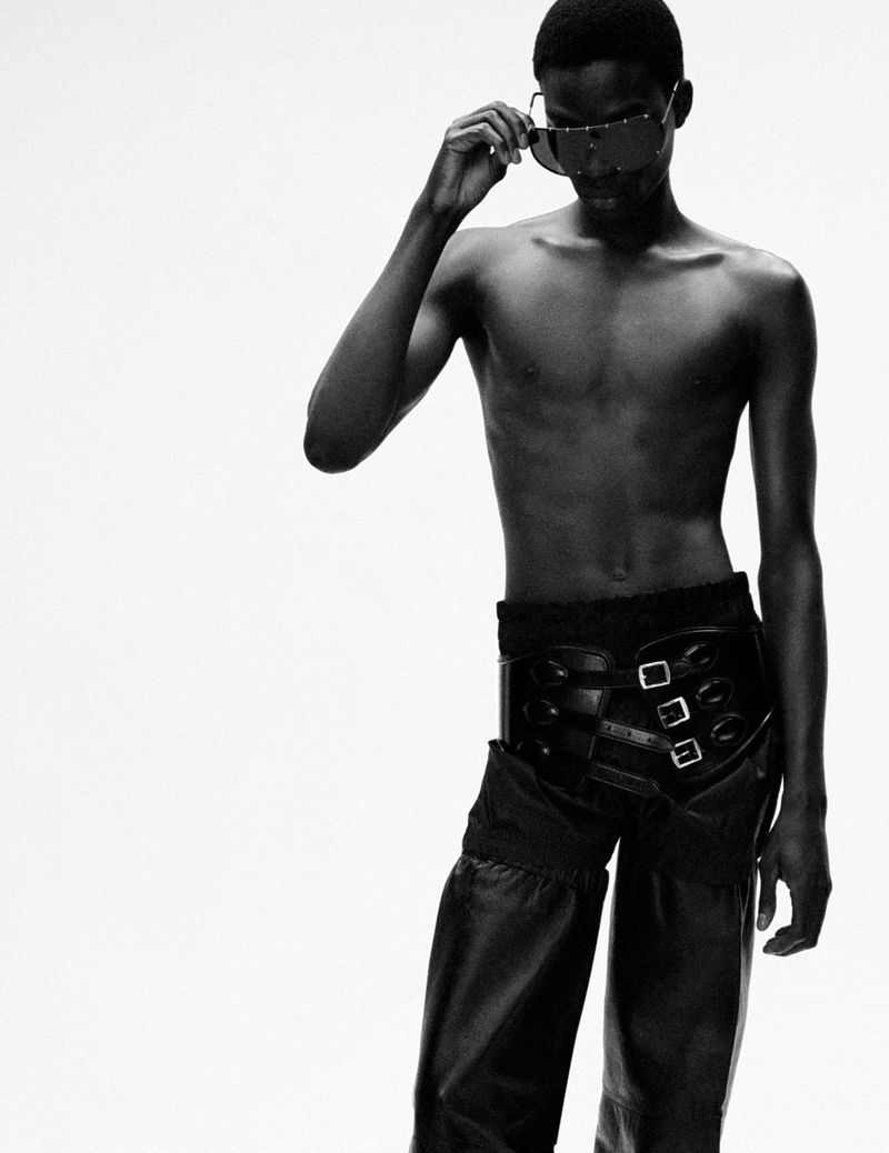 Monochrome studio shot of a shirtless adult male wearing sunglasses and leather chaps standing against a bright white background.
