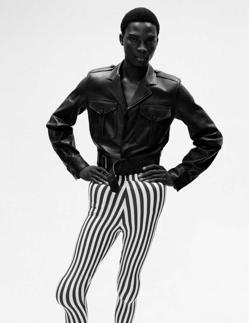 A slim, dark-skinned person with short hair poses in a black leather jacket and striped leggings against a white backdrop in a fashion shot.
