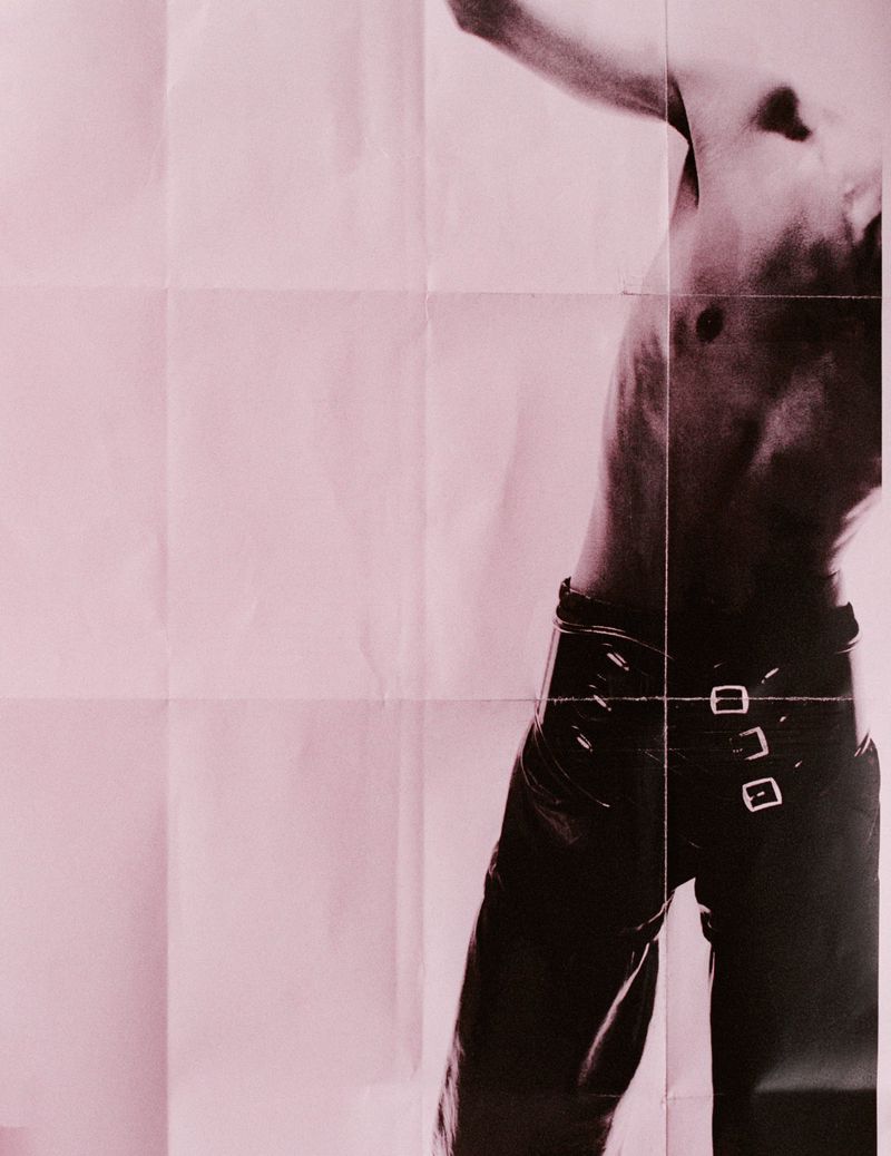 Artistic shot of a shirtless male torso and black leather pants against a pink paper background. Fashion and photographic style.