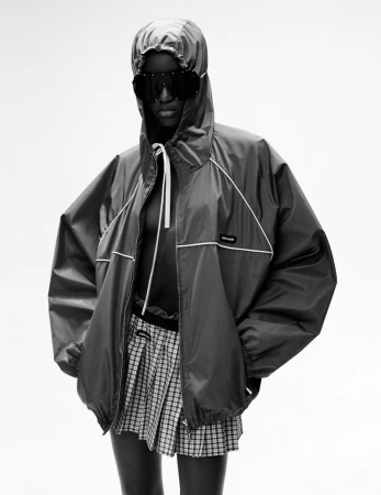 Black and white fashion shot of a dark-skinned person wearing sunglasses, a hooded jacket, and a plaid skirt against a stark white background.