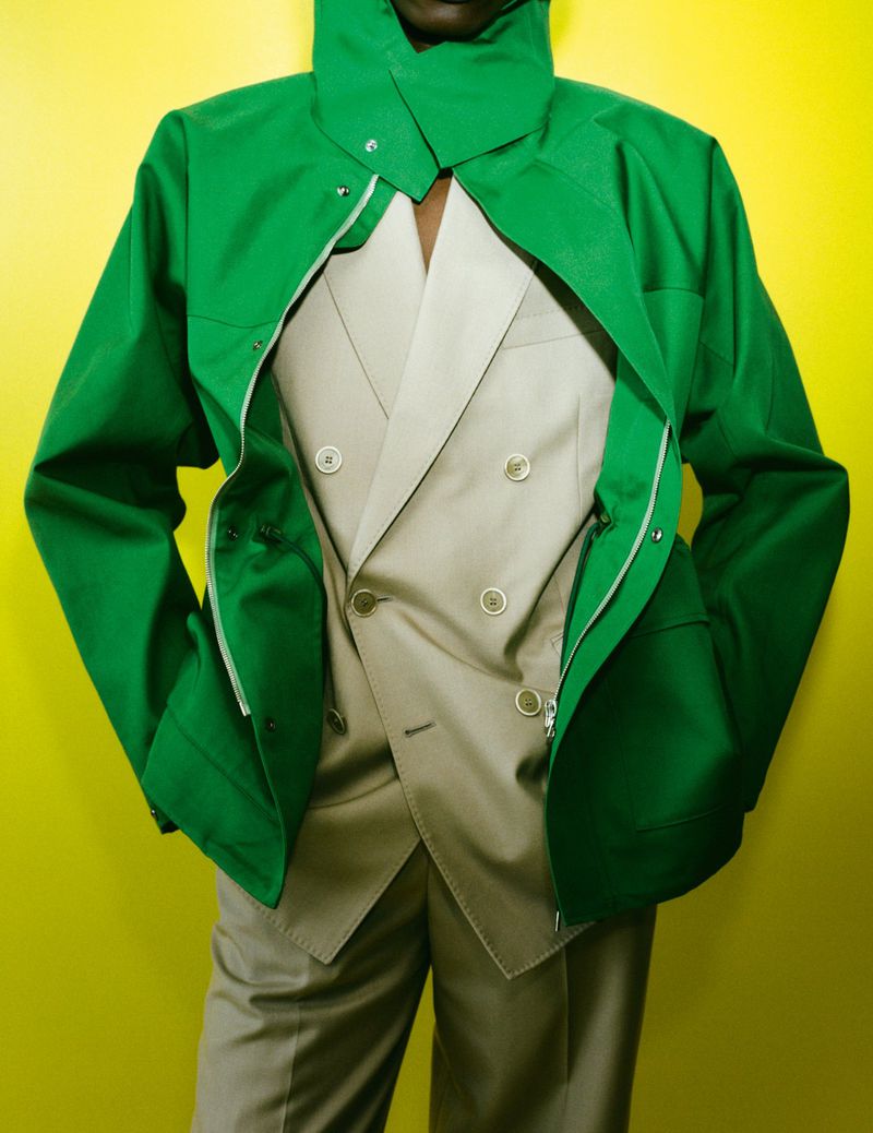 Person in green hooded jacket over khaki suit against bright yellow background. Fashion forward clothing ensemble. 