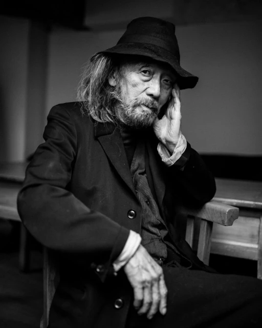 Monochrome portrait of an elderly Asian man with a beard, wearing a dark hat and suit, seated indoors in front of a light wall.