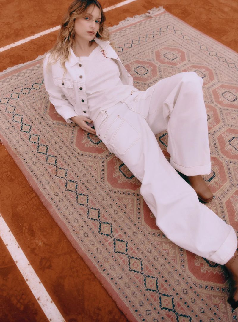 Woman in white denim jacket and jeans lies on patterned rug with brown and white striped floor in background. Fashion editorial.