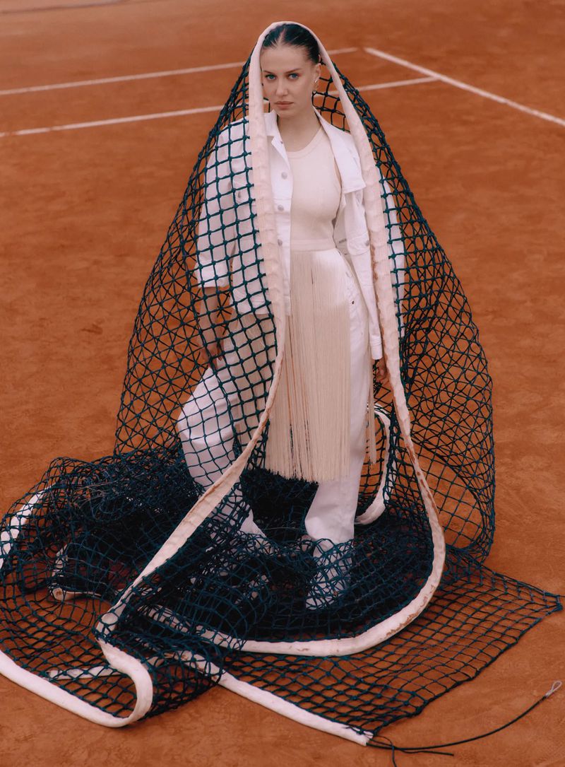 An adult female poses on a tennis court draped in a tennis net, wearing a white jacket and pants with light makeup and hair back.