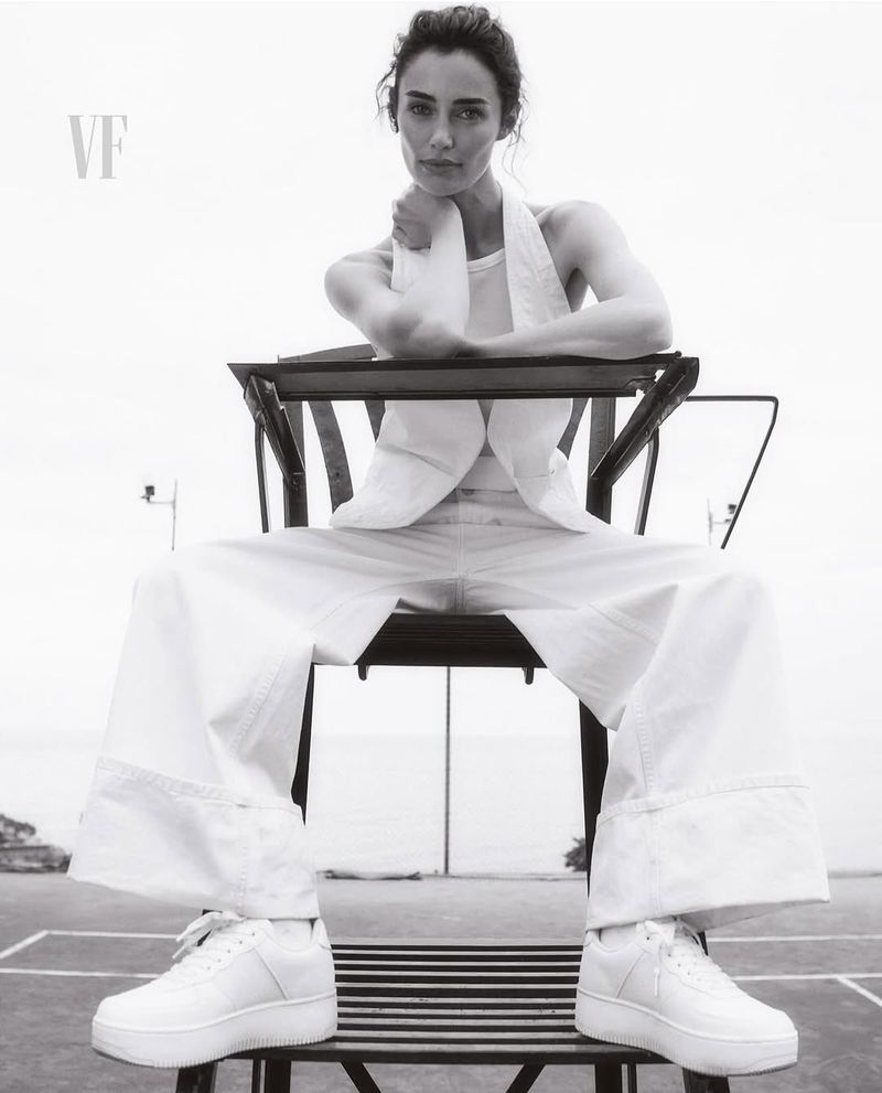 Black and white portrait of a woman in white outfit sitting on umpire chair at a tennis court, looking at the camera with a serious expression.