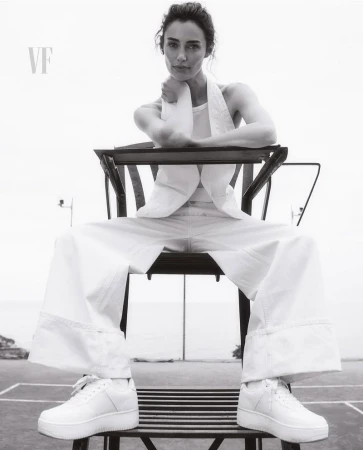 Black and white portrait of a woman in white outfit sitting on umpire chair at a tennis court, looking at the camera with a serious expression.