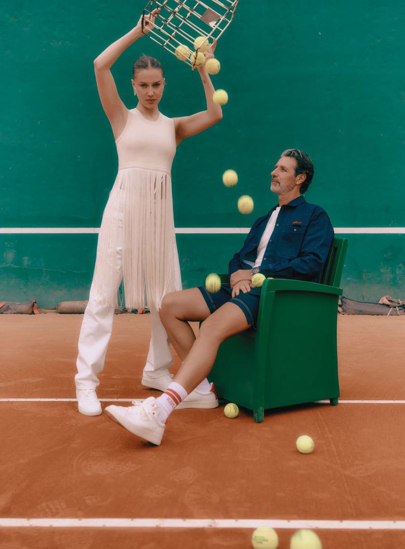 A young woman stands with a tennis ball basket above her head while tennis balls fall, a man sits in a chair in the background.