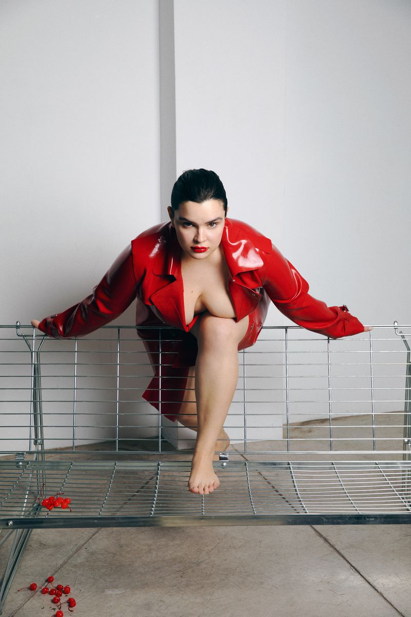 Adult woman in red vinyl jacket crouches on metal grate in indoor photo studio setting with white walls and concrete floor.