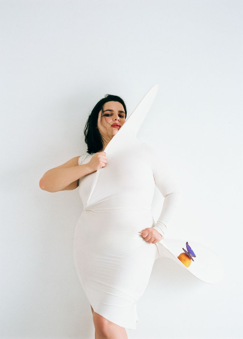 Woman in a white dress holds a large white hat with a flower, posing against a plain white backdrop in a fashion forward style.