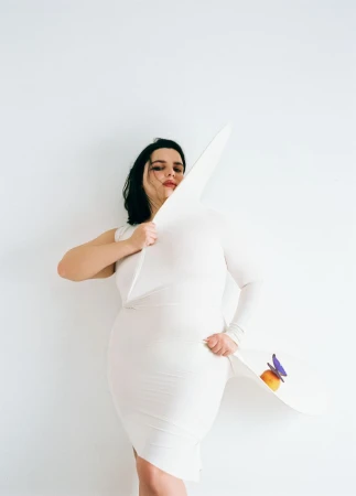 Woman in a white dress holds a large white hat with a flower, posing against a plain white backdrop in a fashion forward style.