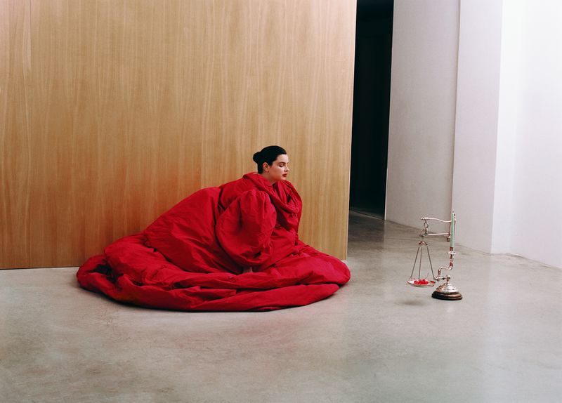 Elegant woman draped in voluminous red fabric, seated on a gray floor in front of a light-wood-paneled wall and scale.