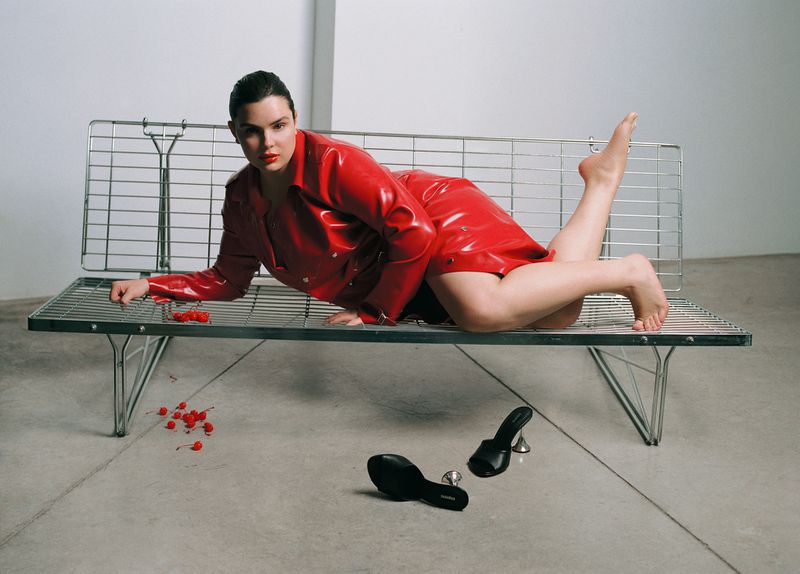 Adult female in a shiny red skirt suit poses on a metal bench indoors next to high-heeled sandals with scattered cherries.