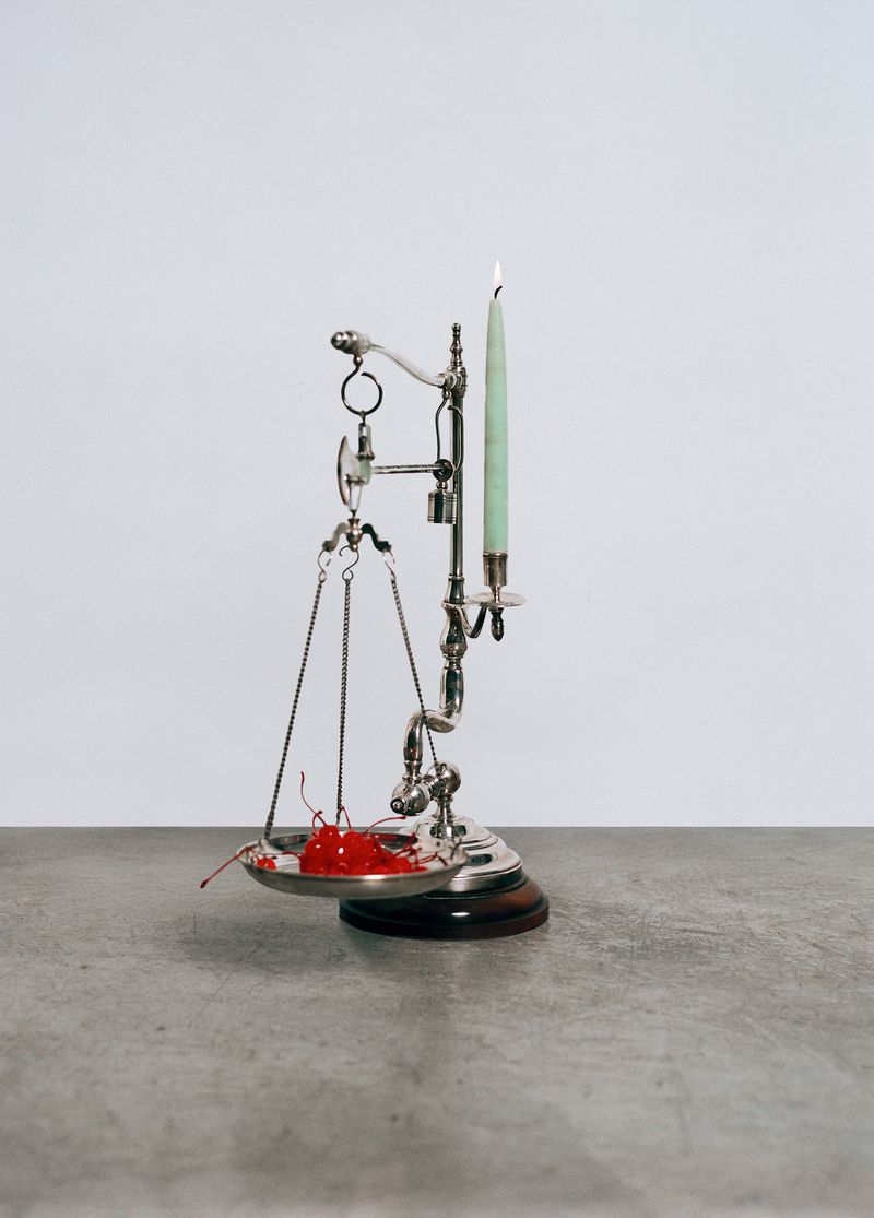 A set of silver scales balances a lit green taper candle on one side with a pile of red cherries on the other against a neutral backdrop.